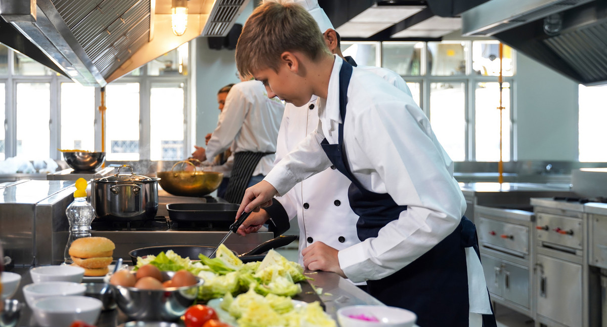 Kochlehrer unterrichtet Schüler in Kochen in einer Küche. Salate und Eier liegen u.a. auf dem Küchentisch.