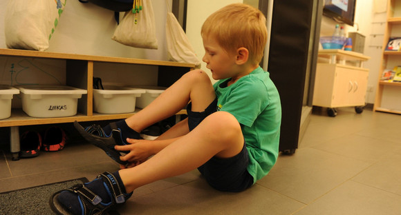 Ein blonder Junge zieht sich seine Schuhe an, hinter ihm hängen an der Wand viele Jacken. 