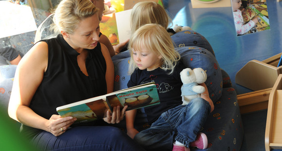 Eine blonde Frau liest einem Mädchen aus einem Bilderbuch vor, beide sitzen auf einem Sofa.  