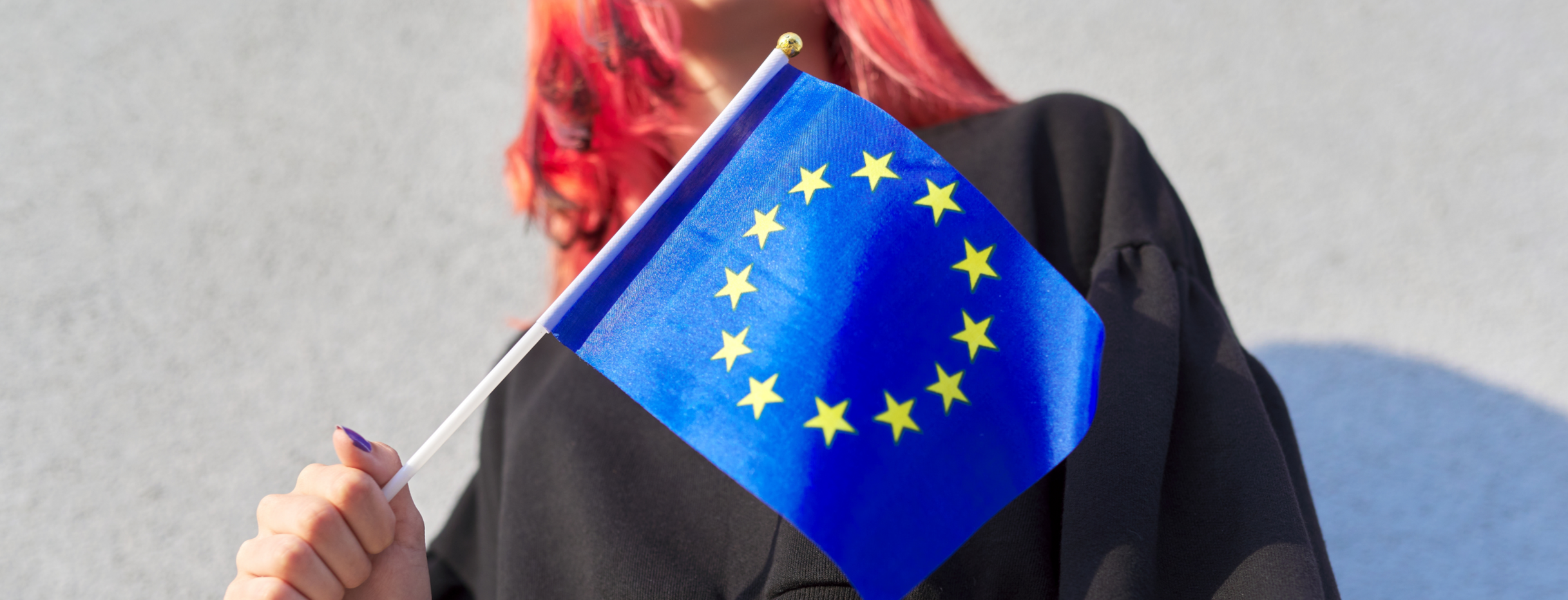 Teilansicht einer jungen Frau mit rotgefärbten Haaren, die eine kleine Europaflagge in der rechten Hand hält.