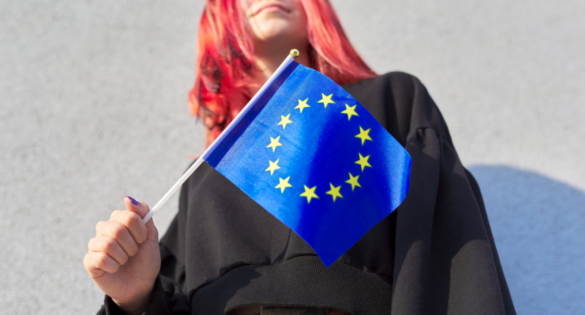 Teilansicht einer jungen Frau mit rotgefärbten Haaren, die eine kleine Europaflagge in der rechten Hand hält.