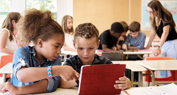 Schulklasse, im Vordergrund eine schwarze Schülerin, die einem ihr neben ihr sitzendem Schüler etwas auf dem Tablet zeigt. Hinten rechts im Hintergrund die stehende Lehrerin.