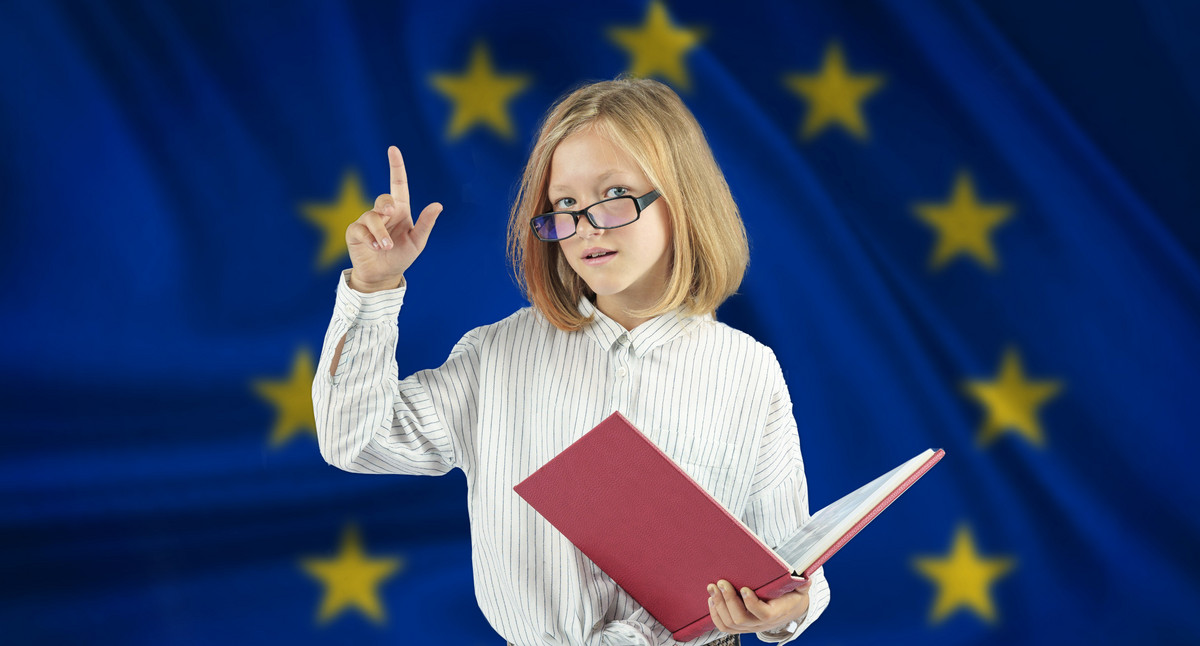 Schülerin mit Brille vor Europaflagge die um Aufmerksamkeit bittent einen Finger hochhält während sie in der anderen Hand ein geöffnetes rotes Buch hält.