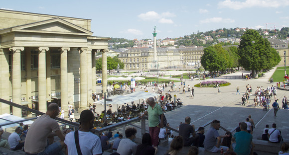 Der Schlossplatz in Stuttgart, aufgenommen von der Treppe, die zum kleinen Schlossplatz hochführt. Viele Menschen sind auf der Treppe und dem Schlossplatz zu sehen.