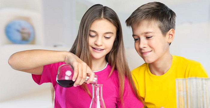 Im Chemieunterricht füllt ein Mädchen in einem roten T-Shirt eine Flüssigkeit in ein Reagenzglas. EIn Junge mit  gelbem T-Shirt schaut zu.