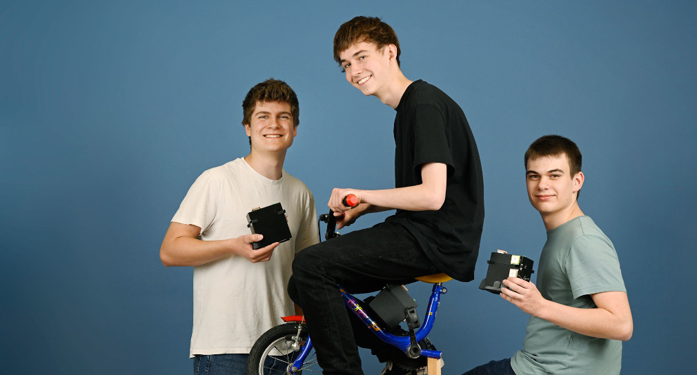 Drei junge Männer vor blauem Hintergrund. Der Junge in der Mitte sitzt auf einem Fahrrad.