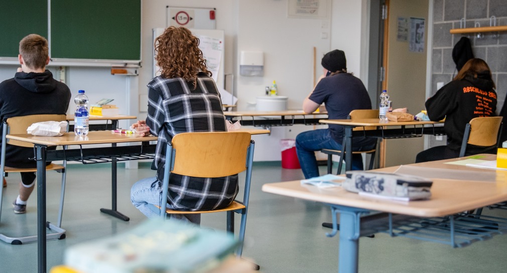 Schülerinnen und Schüler sitzen an Einzeltischen in einem Klassenzimmer, mit dem Rücken zur Kamera. 