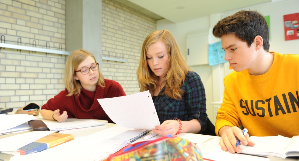 Drei Schüler sitzen an einem Tisch, ein Mädchen hält ein Blatt Papier in der Hand. 