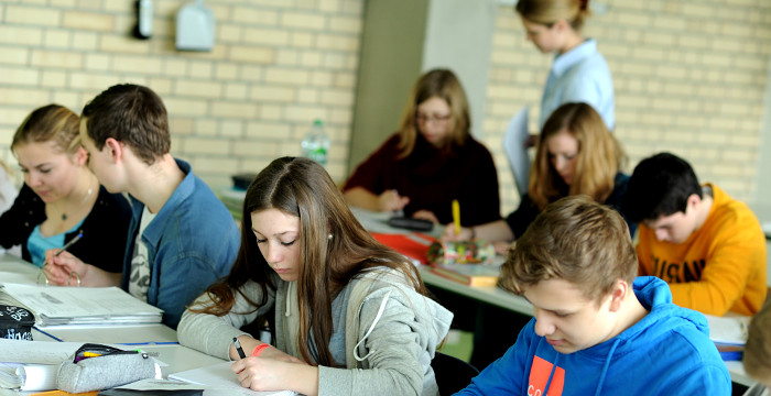 Schülerinnen und Schüler arbeiten im Klassenzimmer am Tisch, während die Lehrerin im Hintergrund herumläuft.