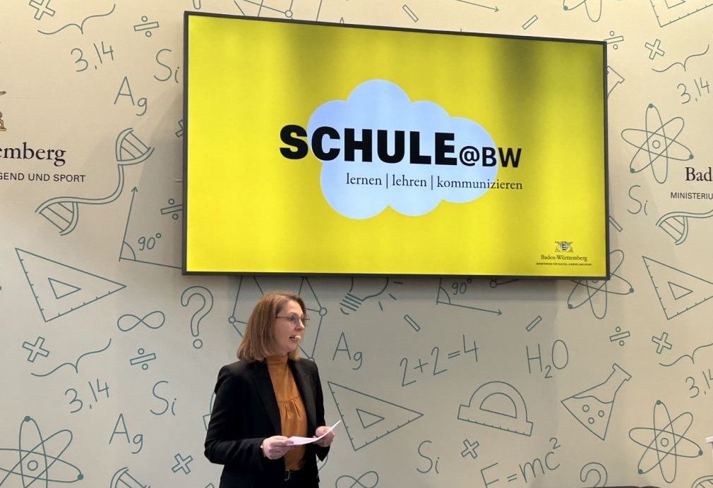 Eine Frau steht vor einer Wand, auf der auf einem großen Bildschirm eine gelbe Schrift zu sehen ist. 