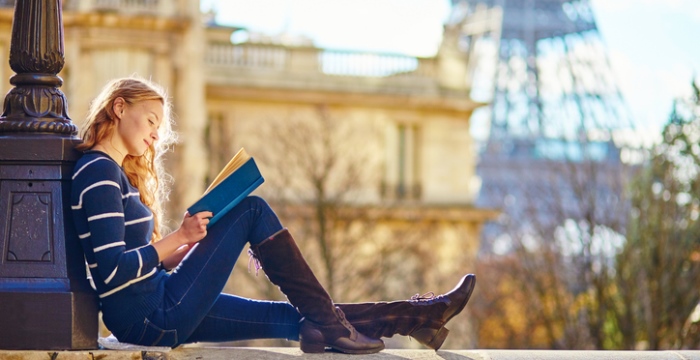 Eine junge Frau sitzt auf einer Mauer und liest ein Buch. Im Hintergrund ist der Eiffelturm zu sehen.