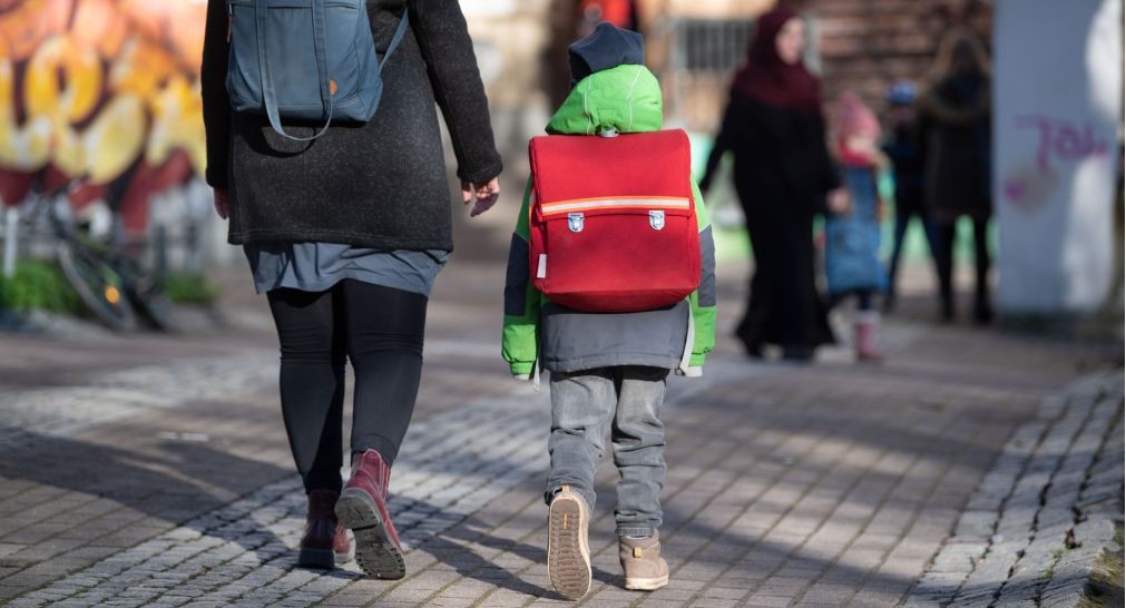 Ein Kind mit rotem Schulranzen läuft neben einer Frau, beide sieht man von hinten. . 