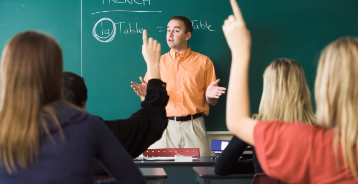 Ein Lehrer steht vor einer Tafel. Mehrere Schülerinnen sitzen im Klassenzimmer und melden sich.
