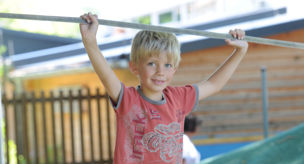 Ein Junge mit blauen Augen und blonden Haar hält sich an einem Seil fest und lächelt in die Kamera