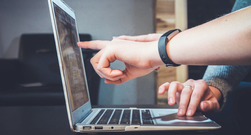 Zu sehen ist eine Hand, die auf den Bildschirm eines Laptops zeigt. Die Hand einer anderen Person bedient das Laptop.