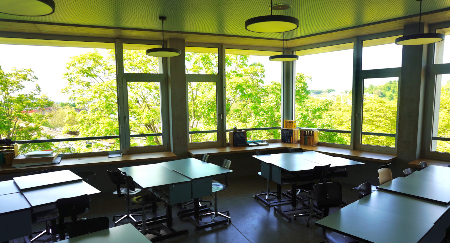 Ein leerer Klassenraum mit mehreren Tischen und Stühlen drin, durch die Fenster sieht man Bäume. 