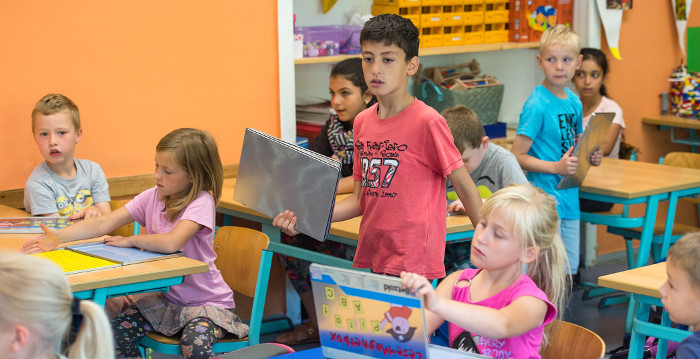 Ein Junge mit  dunkler Hautfarbe geht im Klassenzimmer zwischen den Stühlen nach vorne, in seiner Hand hält er einen graue Mappe.