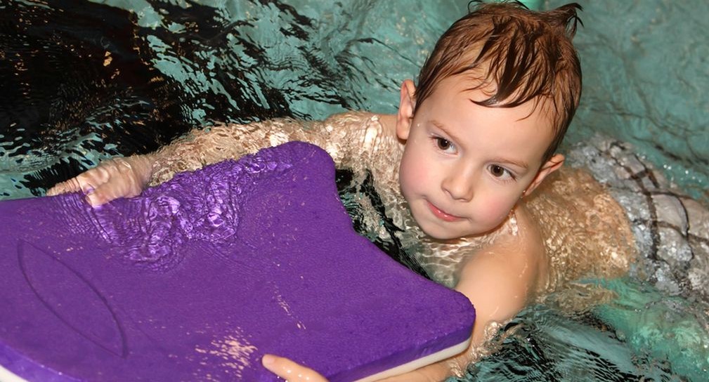 Ein Kind schwimmt im Wasser und hat eine lilanes Schwimmbrett in der Hand. 