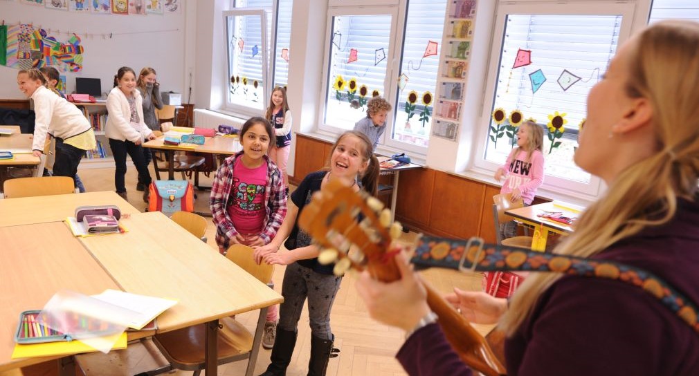 Kinder singen im Klassenzimmer, eine Frau spielt dazu Gitarre.