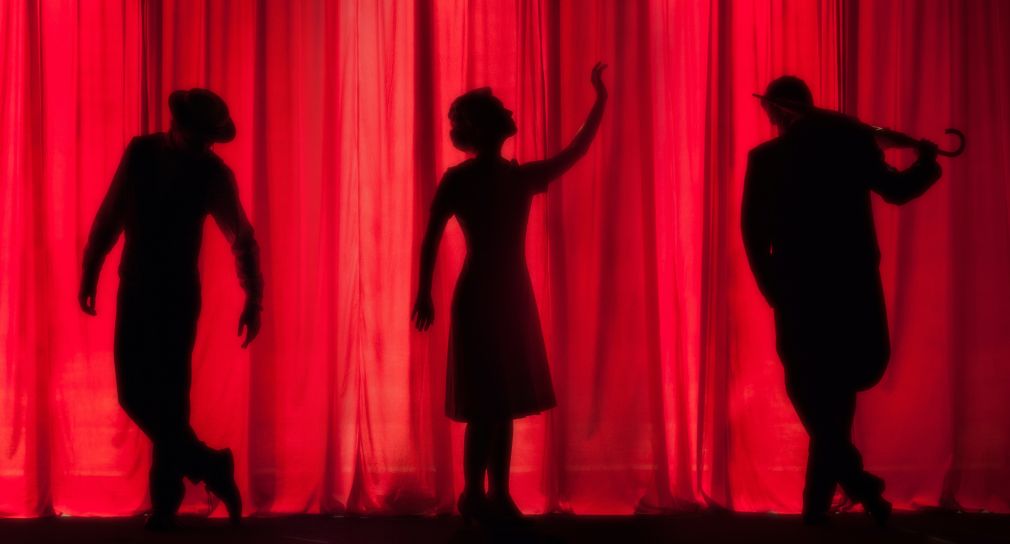 Die Silhouetten von zwei Schauspielern und einer Schauspielerin vor einem roten Vorhang