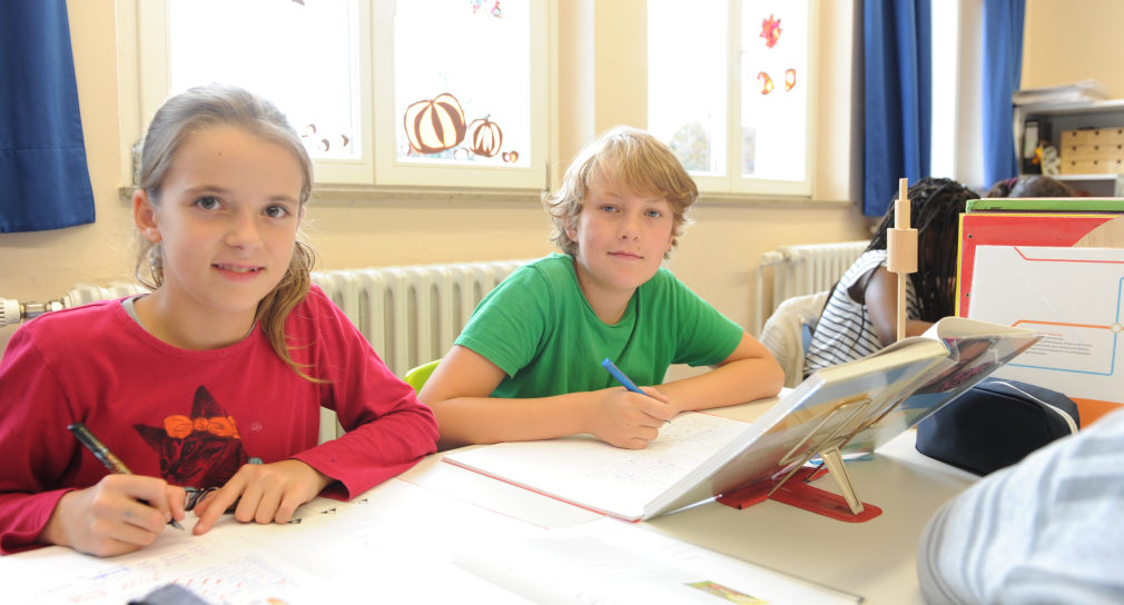 Ein Mädchen und ein Junge sitzen nebeneinander in einem Schulraum. Vor ihnen liegen aufgeschlagene Hefte und Bücher