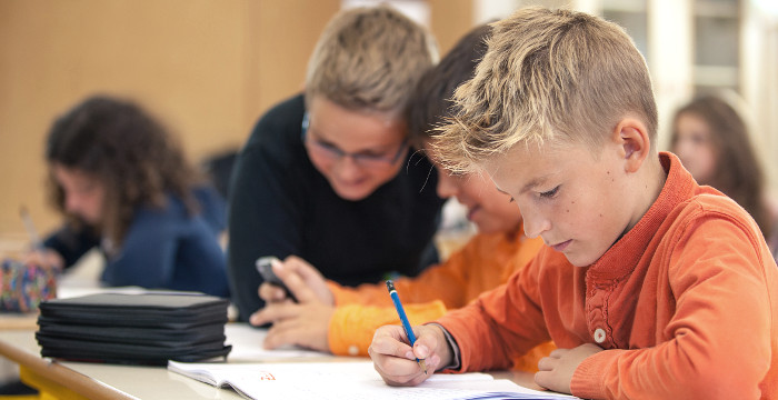Ein Junge mit orangenem Pullover schreibt am Schultisch.