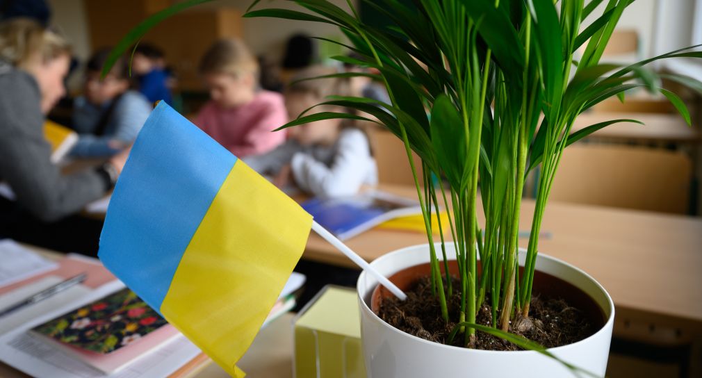 Grundschulkinder sitzen in einem Klassenzimmer. Im Vordergrund ist ein ukrainisches Fähnchen in einem Blumentopf.