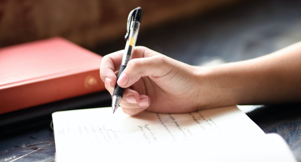 Abgebildet ist eine Hand, die mit einem Stift in ein Notizbuch schreibt.