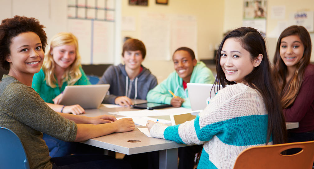 Sechs Schülerinnen und Schüler sitzen gemeinsam an einem Gruppentisch und lächeln in die Kamera.