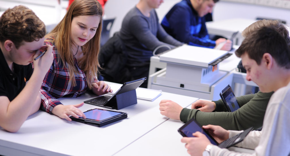 Mehrere Schüler und eine Schülerin sitzen mit Tablets an Tischen in einem Unterrichtsraum